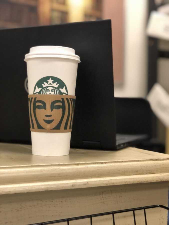 A Starbucks coffee cup sits on a desk with a laptop directly behind it. I love going to Starbucks before school because I can enjoy  it at class, said Jade Galan.